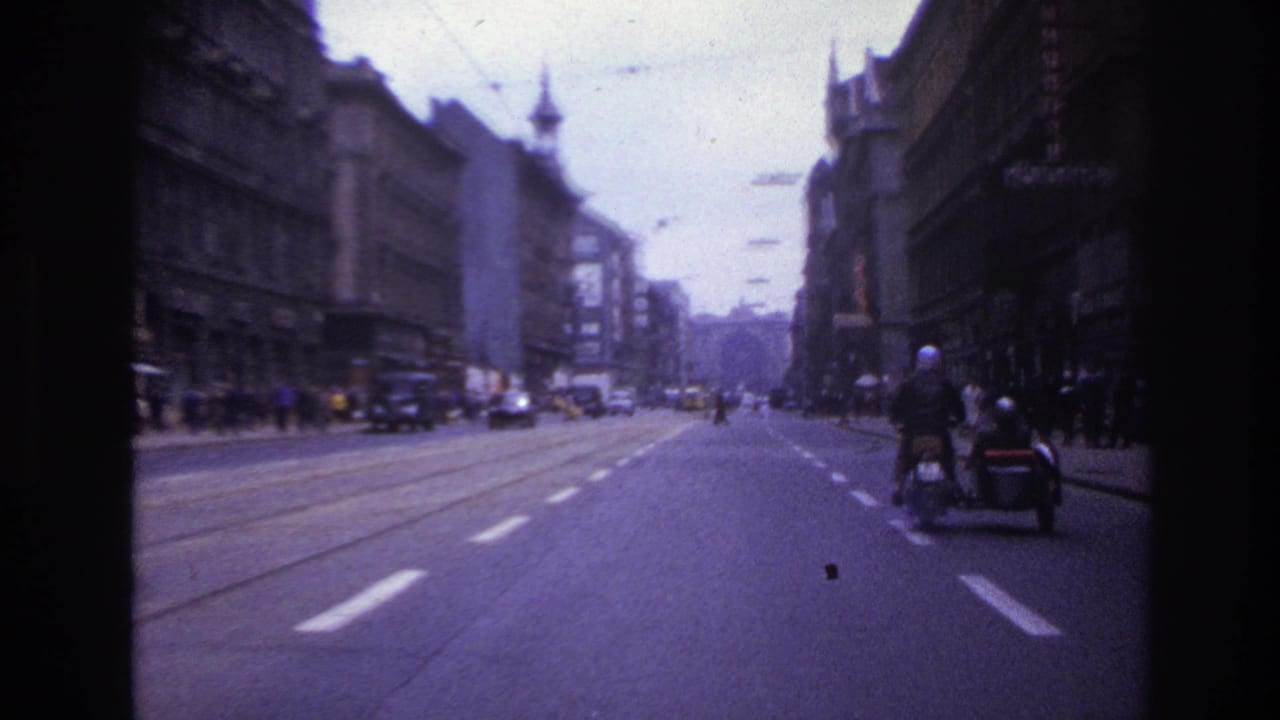 Broad city road with tram tracks, cars, sidewalks, and a motorcycle with sidecar moving ahead on the right.