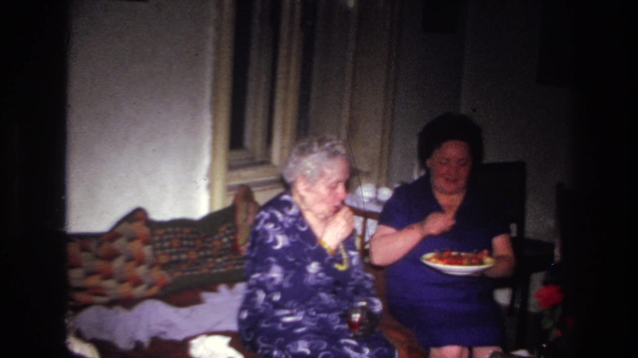 Two older women sit in a private interior in France, 1967, with one eating fruit from a plate beside a bed or daybed.