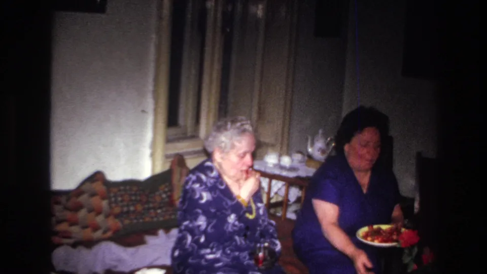 In a 1967 France home-movie interior, two older women sit near a bed or daybed while a plate of fruit is held in the foreground.