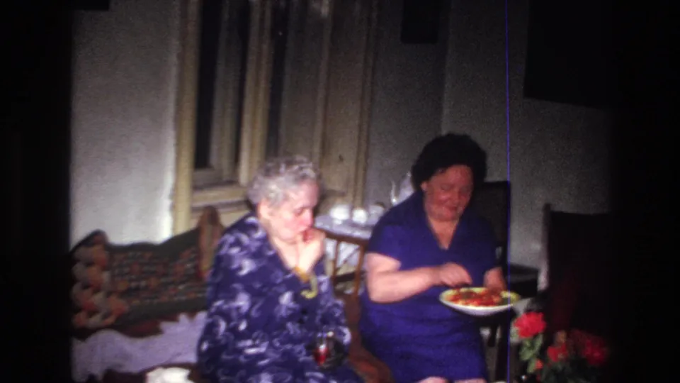 Two older women in a 1967 France home-movie interior sit together as one holds a fruit plate near a small red flower arrangement.