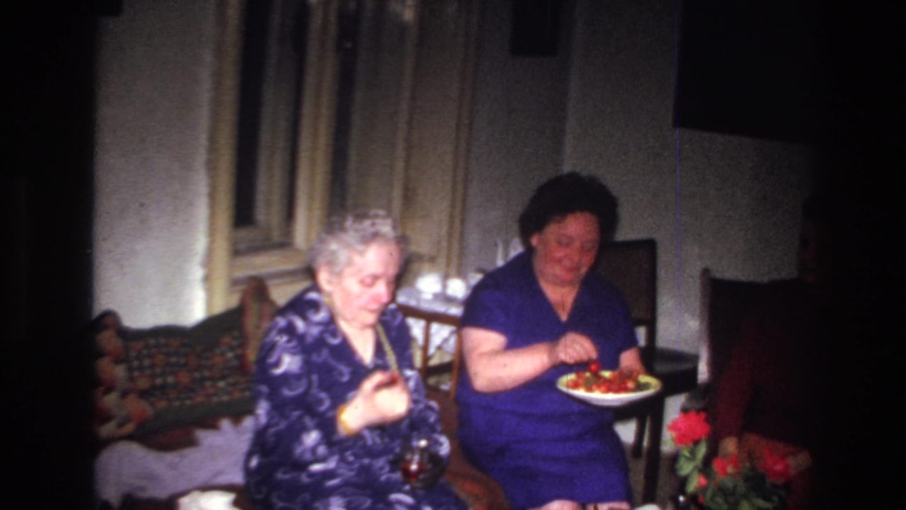 In 1967 France home-movie footage, two women sit near a patterned couch as one holds a glass and the other eats from a plate.