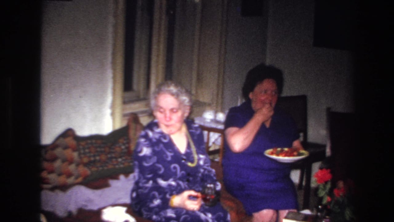 Two women seated by a couch, with one woman holding a plate and raising food to her mouth.