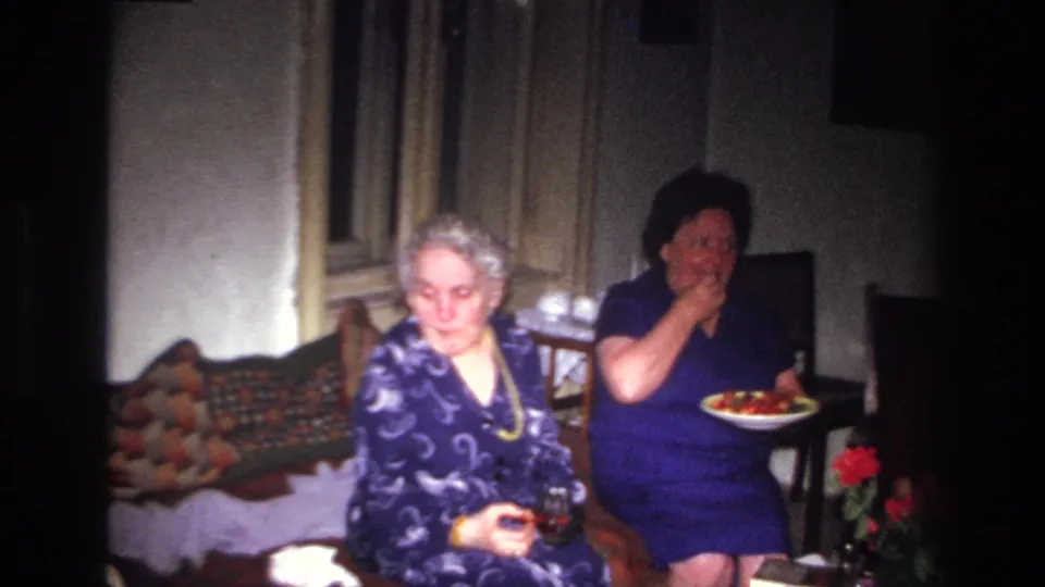 In the French 1967 interior scene, one woman holds a plate of food while the white-haired woman sits beside her with a glass.