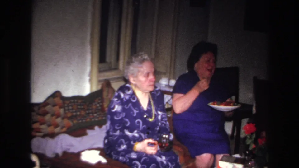 1967 France home-movie frame of two women on chairs near a couch, with one eating and appearing to laugh or speak.