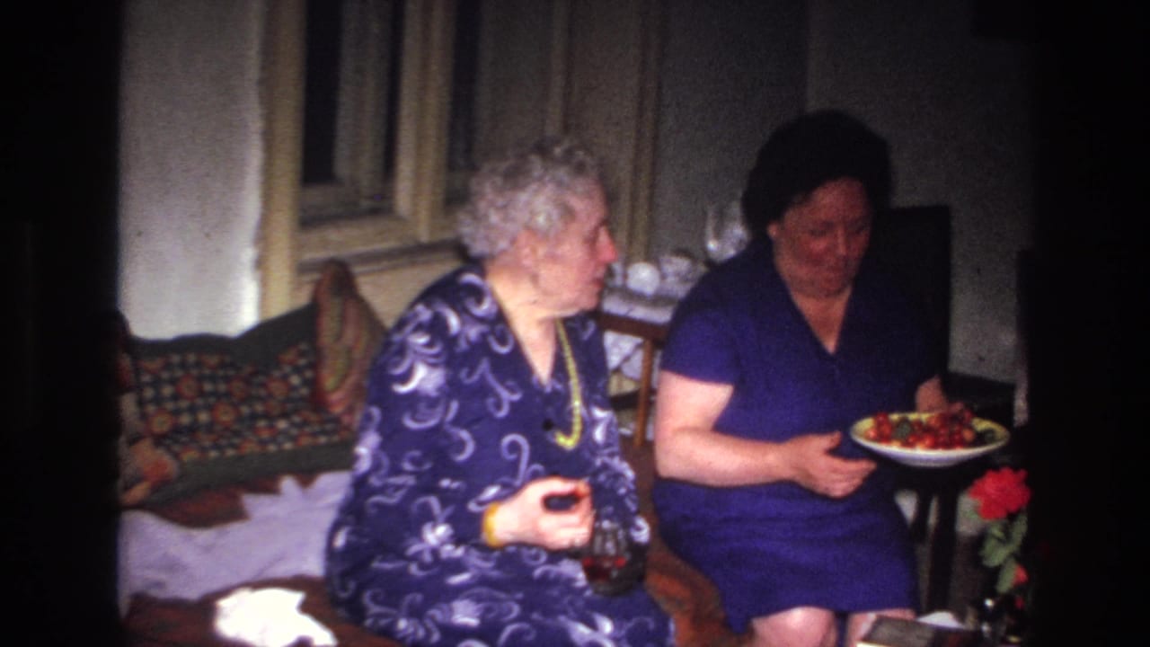 In 1967 France home-movie footage, two older women sit in a dim room with patterned cushions, a glass, and a plate of food.