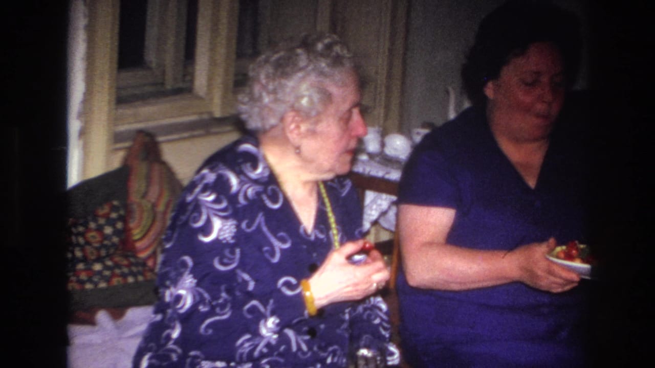 An older woman in a patterned dress turns toward another woman holding a plate of food.