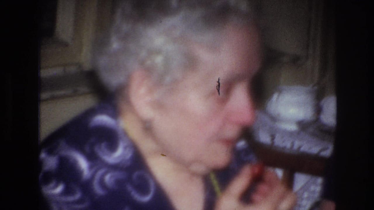 1967 France home-movie frame of an elderly woman in a purple patterned shirt and yellow beads holding food in a domestic interior.