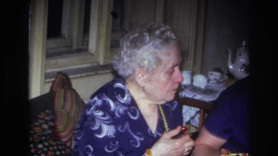 1967 French home-movie frame showing the elderly woman seated with food beside another partially visible woman in a private interior.