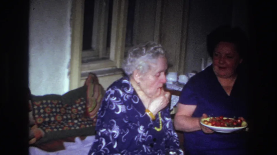1967 France home-movie frame of two women seated indoors, with the elderly woman eating and another woman holding a plate of red snacks.