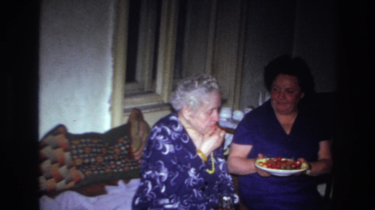 In a 1967 France home-movie frame, an older woman sits on a patterned sofa as a younger woman offers a plate of red fruit.