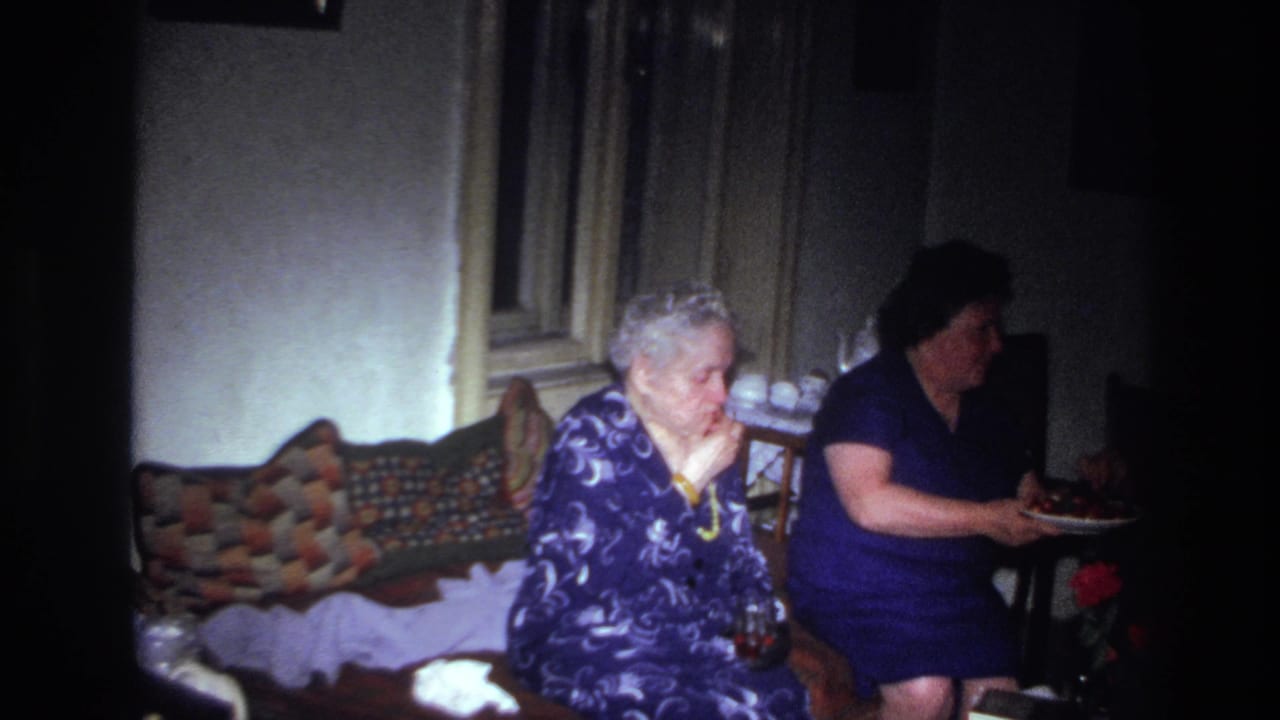 Older woman eating fruit on a sofa as another woman turns away with the plate.
