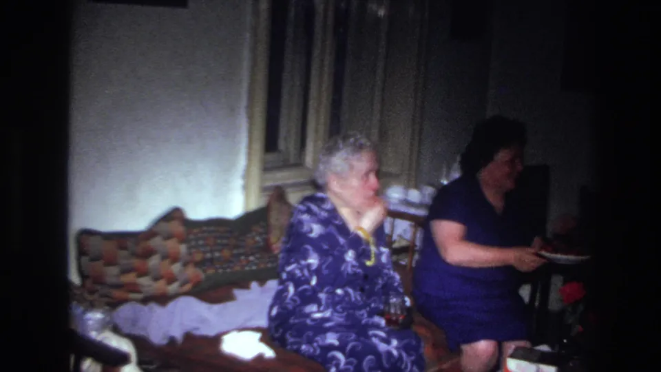 In the dim interior, the older woman sits on a patterned sofa as the younger woman sits beside her with a plate of fruit.