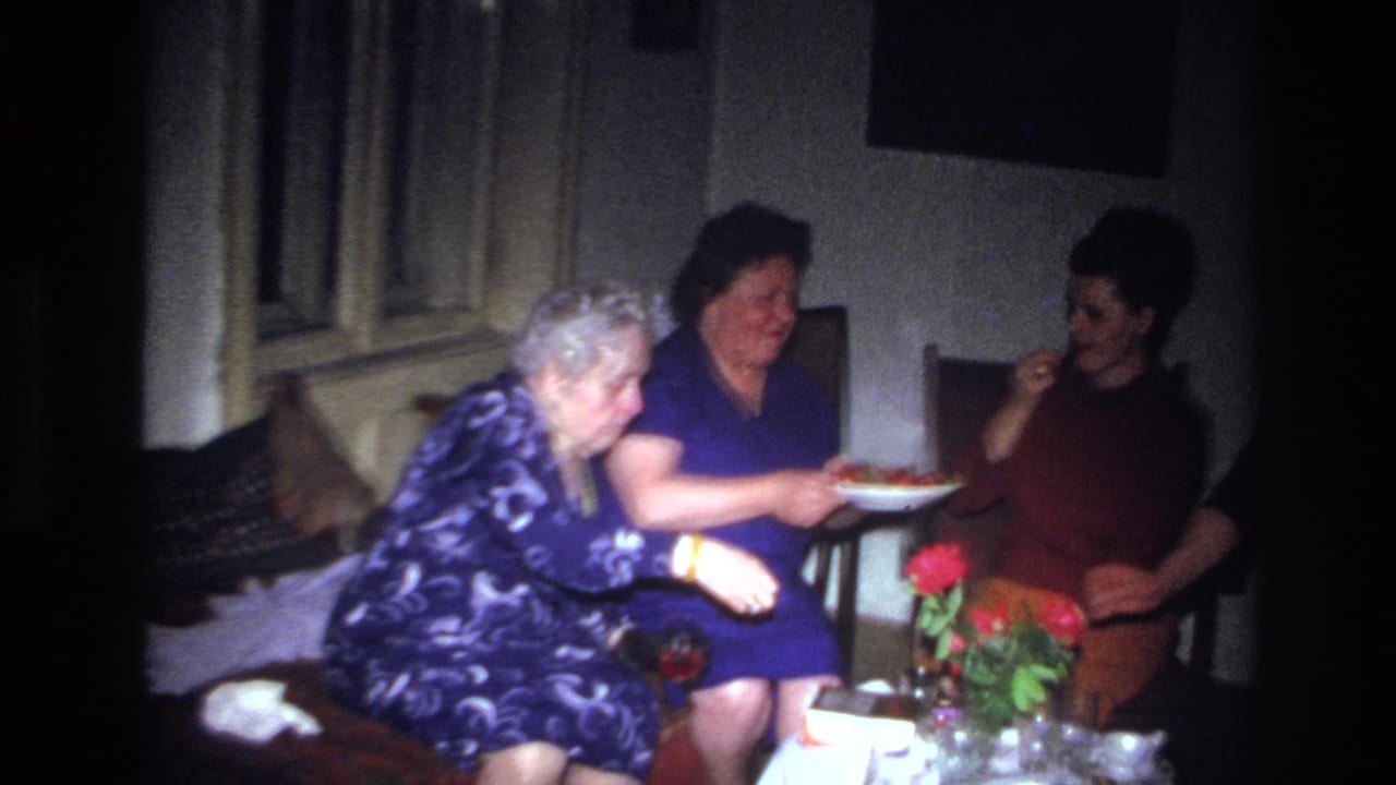 In a dim French home interior from the 1967 8mm clip, three seated women gather near a small table as a woman in a blue dress passes a plate of food.