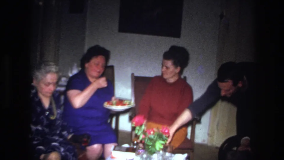 Four adults are visible in the 1967 France home-movie dinner scene, with two women seated centrally, an older woman at left, and a man leaning near a table with red flowers.