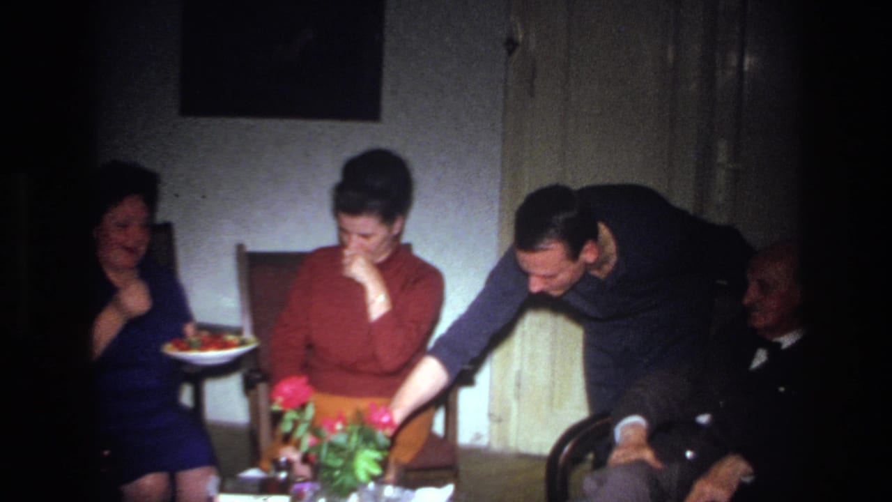 1967 France home-movie frame of adults in a private room, with a younger man leaning over a table beside seated family members.