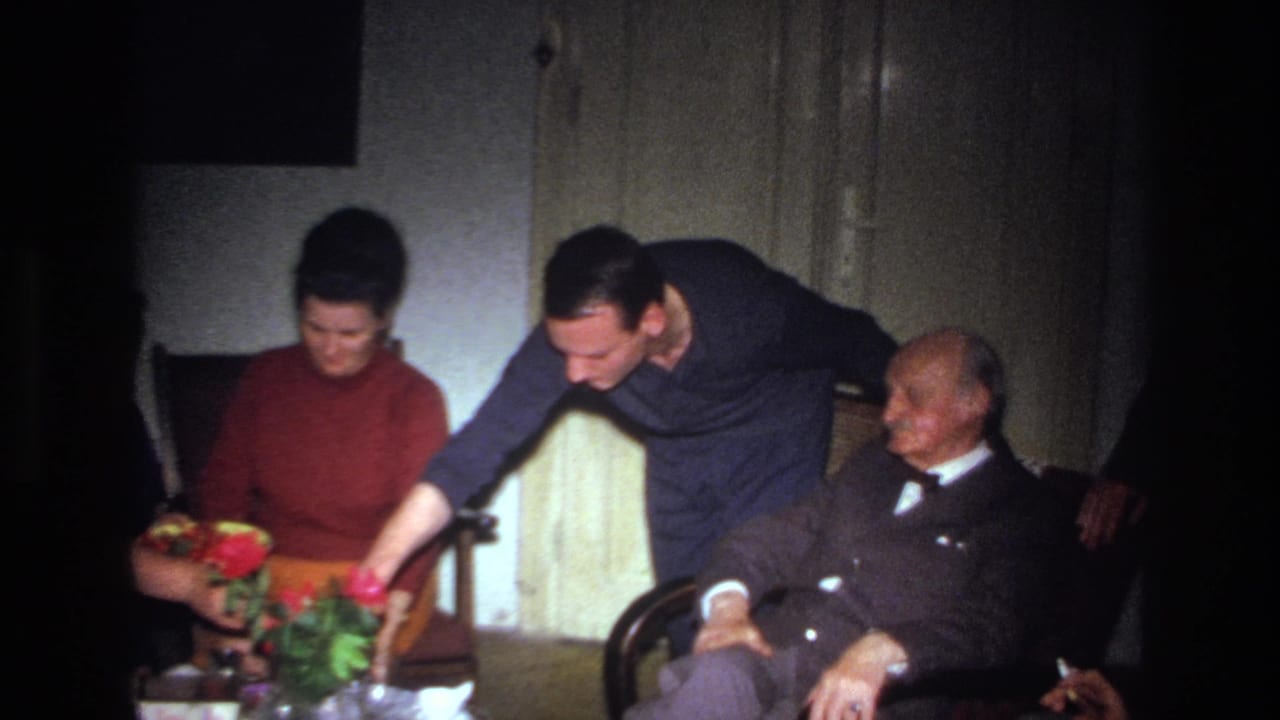 1967 France home-movie frame showing a younger man serving food near a seated woman and an elderly man in a suit and bow tie.