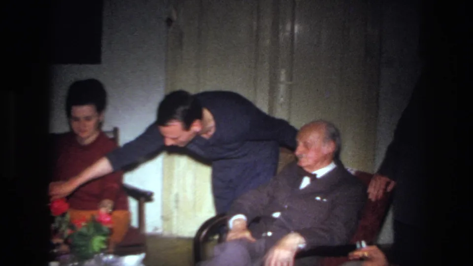 1967 France home-movie frame with a younger man leaning over a flowered table as an elderly seated man and woman look on.