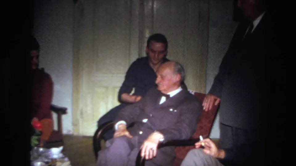 1967 France home-movie frame of an elderly man seated in formal clothing with a younger man behind him and other adults nearby.
