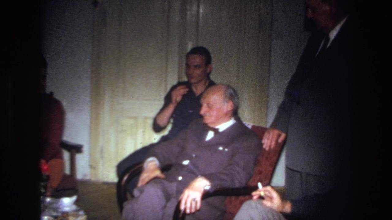 1967 France home-movie frame showing an elderly suited man resting in a red chair while a younger man sits nearby in a dim room.
