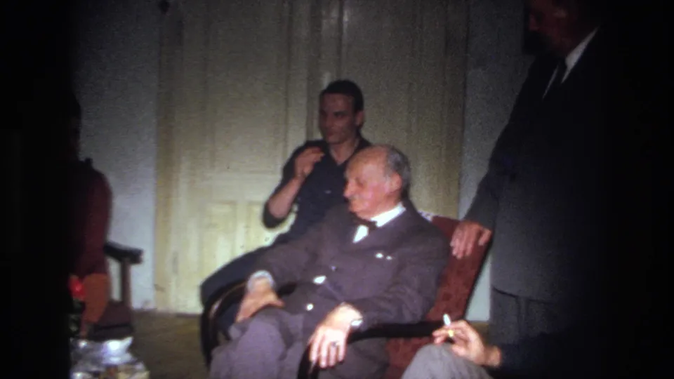 1967 France home-movie frame showing an elderly suited man resting in a red chair while a younger man sits nearby in a dim room.