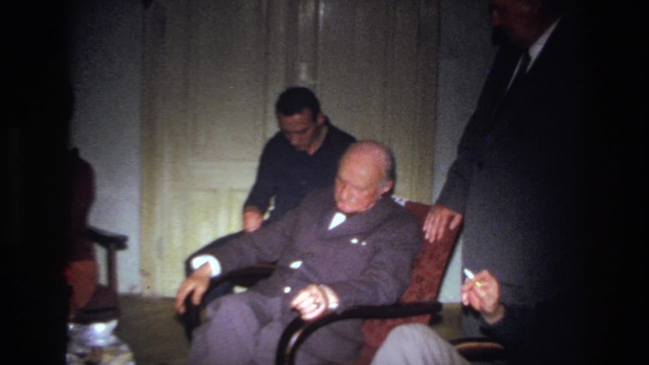 Younger man leans forward behind a seated elderly man in a red chair.
