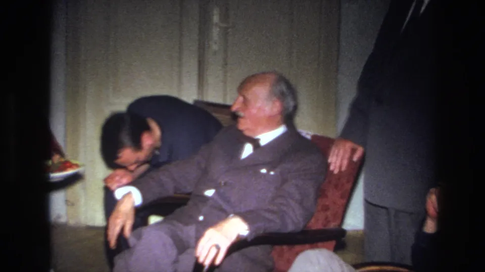The younger man bends down beside the chair as the elderly man turns awake or attentive, surrounded by partial figures in a private room.