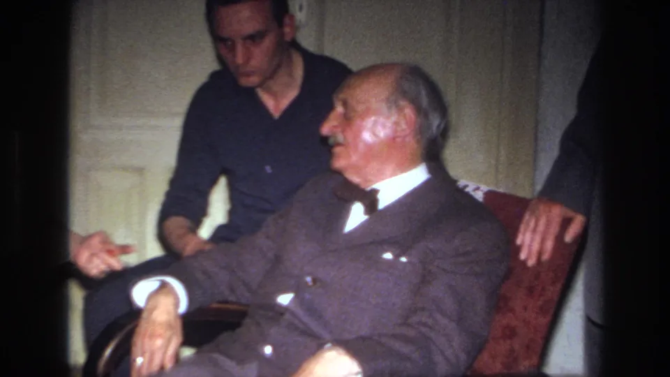 The elderly man appears awake and animated in the red chair while the younger man watches from close behind in the 1967 France interior scene.