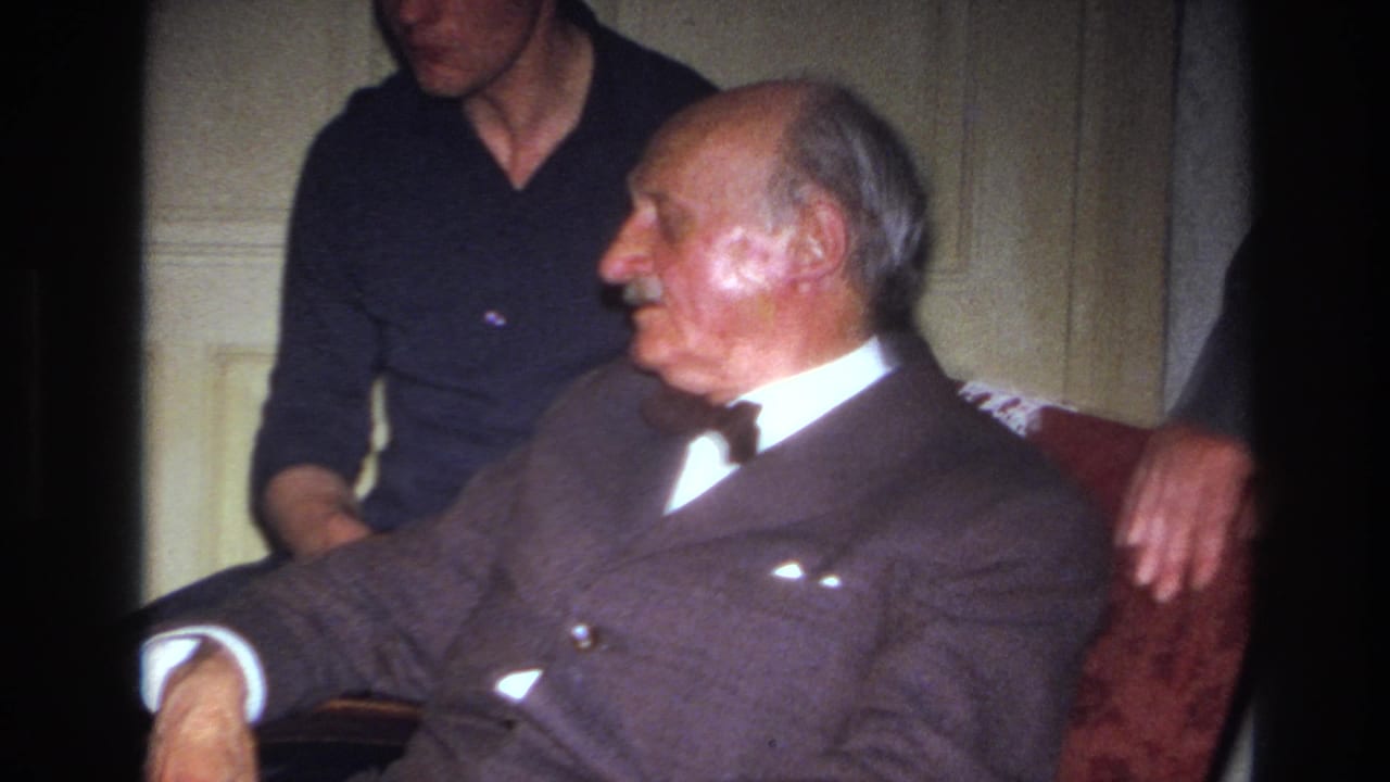 In a 1967 France home-movie frame, an elderly mustached man in a dark suit and bow tie sits on a red sofa while a younger man in a dark sweater stands close behind him.
