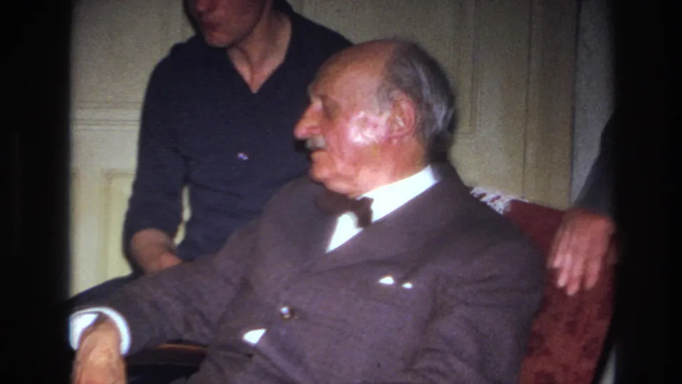 In a 1967 France home-movie frame, an elderly mustached man in a dark suit and bow tie sits on a red sofa while a younger man in a dark sweater stands close behind him.