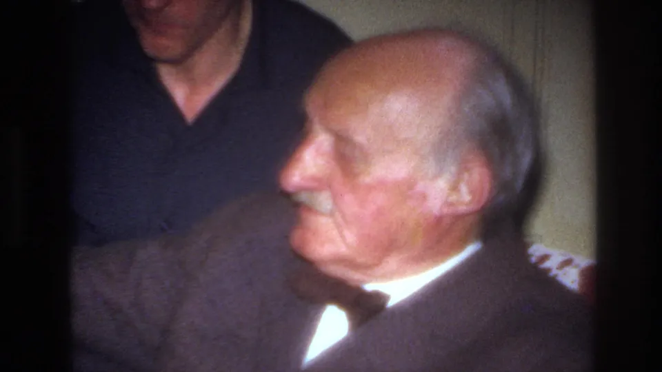 The elderly man is shown in closer profile on the sofa, with his bow tie and suit visible and the younger man partly cropped behind him in the private interior.