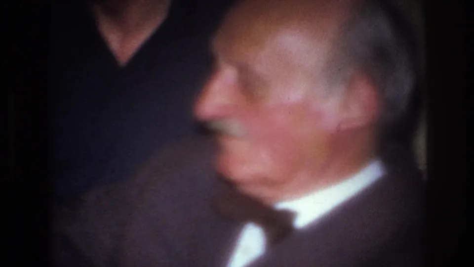 A soft, blurred close-up catches the elderly man's face and suit in motion, preserving the handheld home-movie feel of the living-room scene.