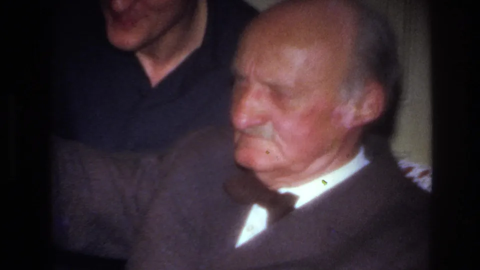 The elderly man remains seated on the sofa in his suit and bow tie while the younger man stands behind him, partly cropped, in the same warmly lit French home interior.