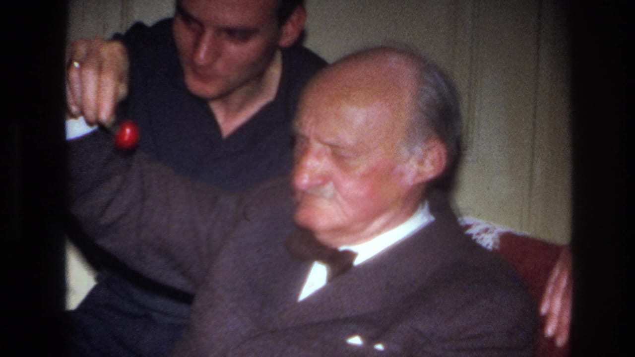 In a 1967 France home-movie frame, a younger man offers a red cherry to an elderly man seated in an armchair.