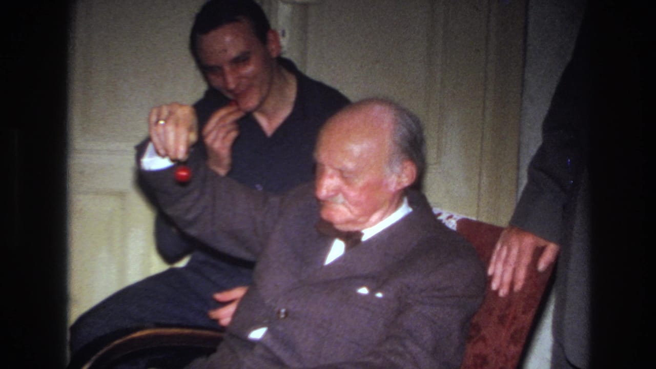 Elderly man sits in profile as a younger man smiles and holds a cherry in front of him.