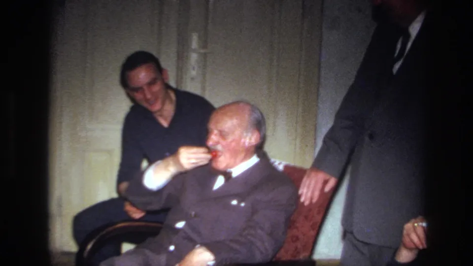 Inside the room, the younger man places a cherry at the elderly man's mouth as a suited man stands beside the chair.