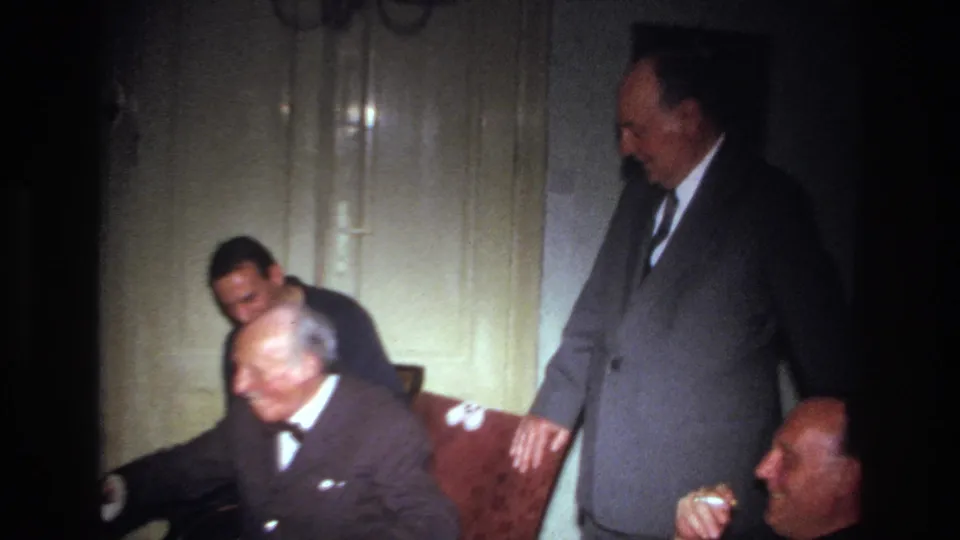 The elderly man leans forward in the armchair while a suited man and the younger man remain close in the dim interior.
