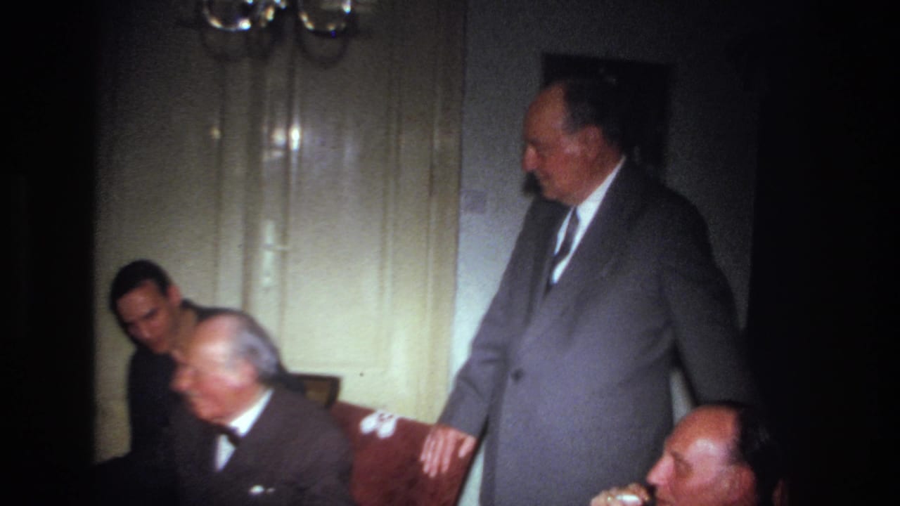 1967 France home-movie frame showing adult men in suits gathered indoors, with one man standing beside seated guests.