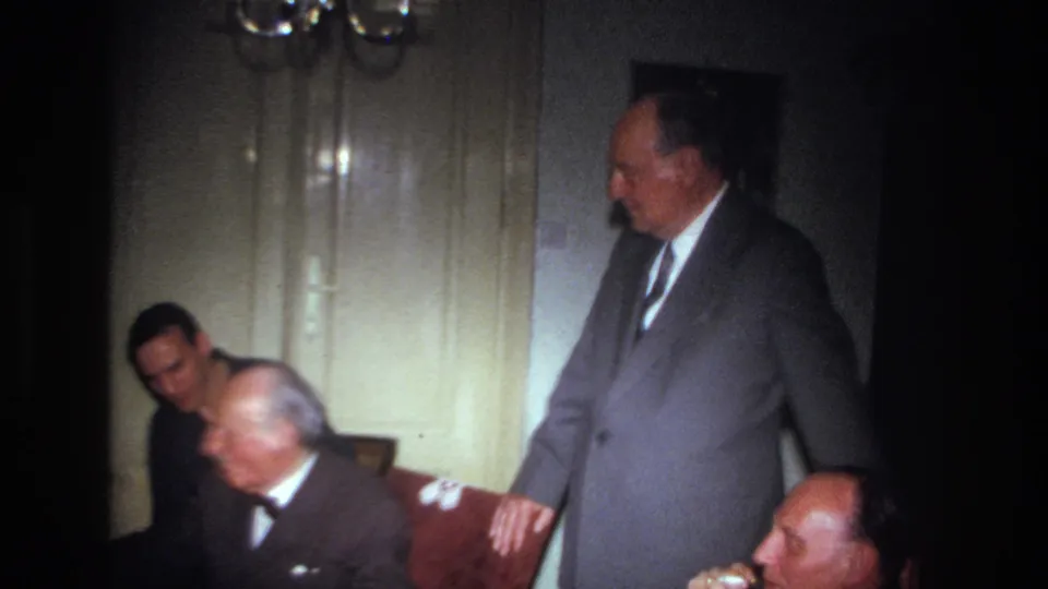 1967 France home-movie frame showing adult men in suits gathered indoors, with one man standing beside seated guests.