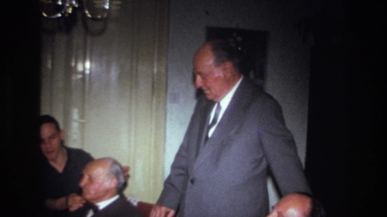 1967 France home-movie frame of a suited man standing near seated guests in a private interior with pale doors and chandelier light.
