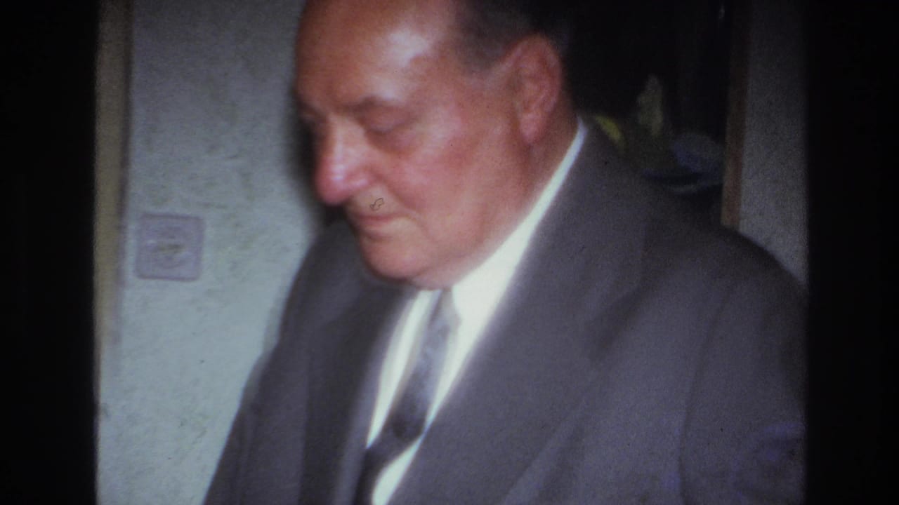 In 1967 France home-movie footage, an older man in a gray suit and tie looks downward inside a softly lit private room.