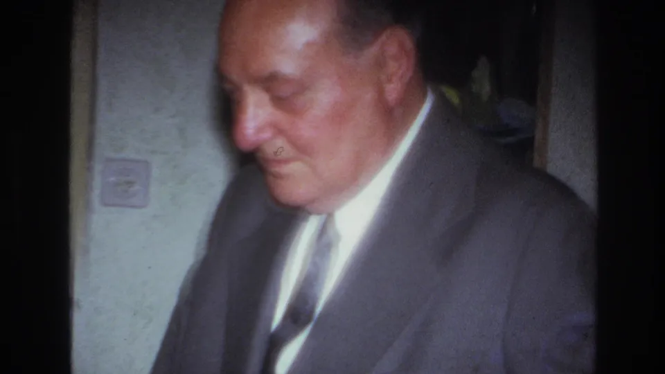 In 1967 France home-movie footage, an older man in a gray suit and tie looks downward inside a softly lit private room.