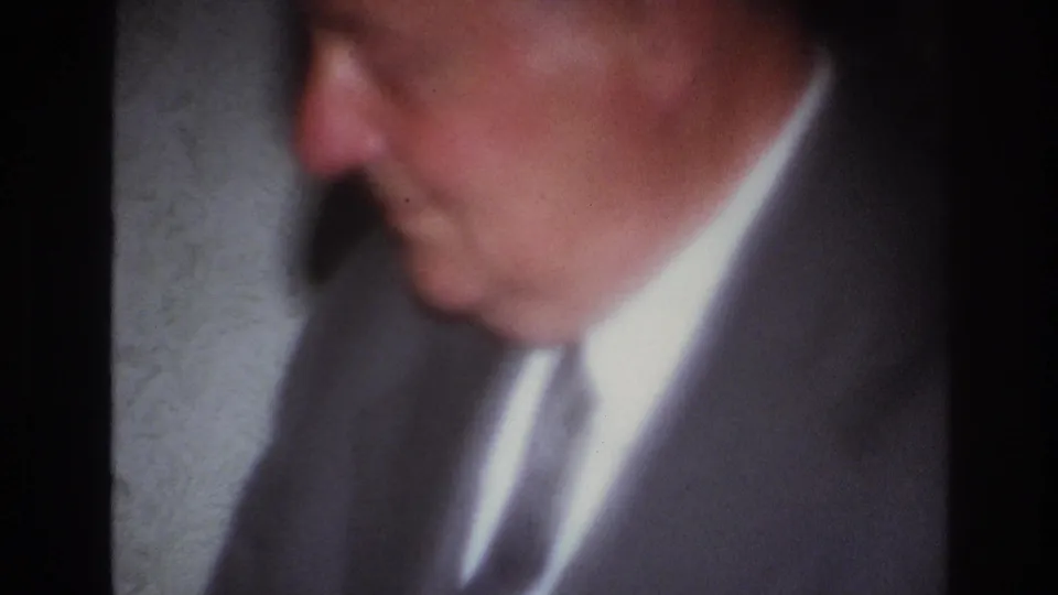 A soft, close 8mm view shows a suited older man turned in profile within the dimly lit French interior.
