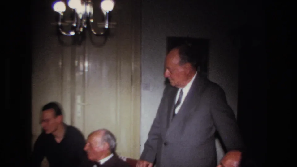 Several men appear in a warm private interior as one suited older man stands near seated guests beneath a chandelier.