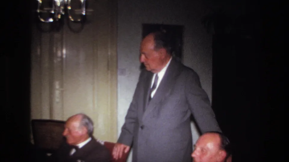 The suited older man stands beside seated men in a dim French private room with a chandelier and pale doors behind them.