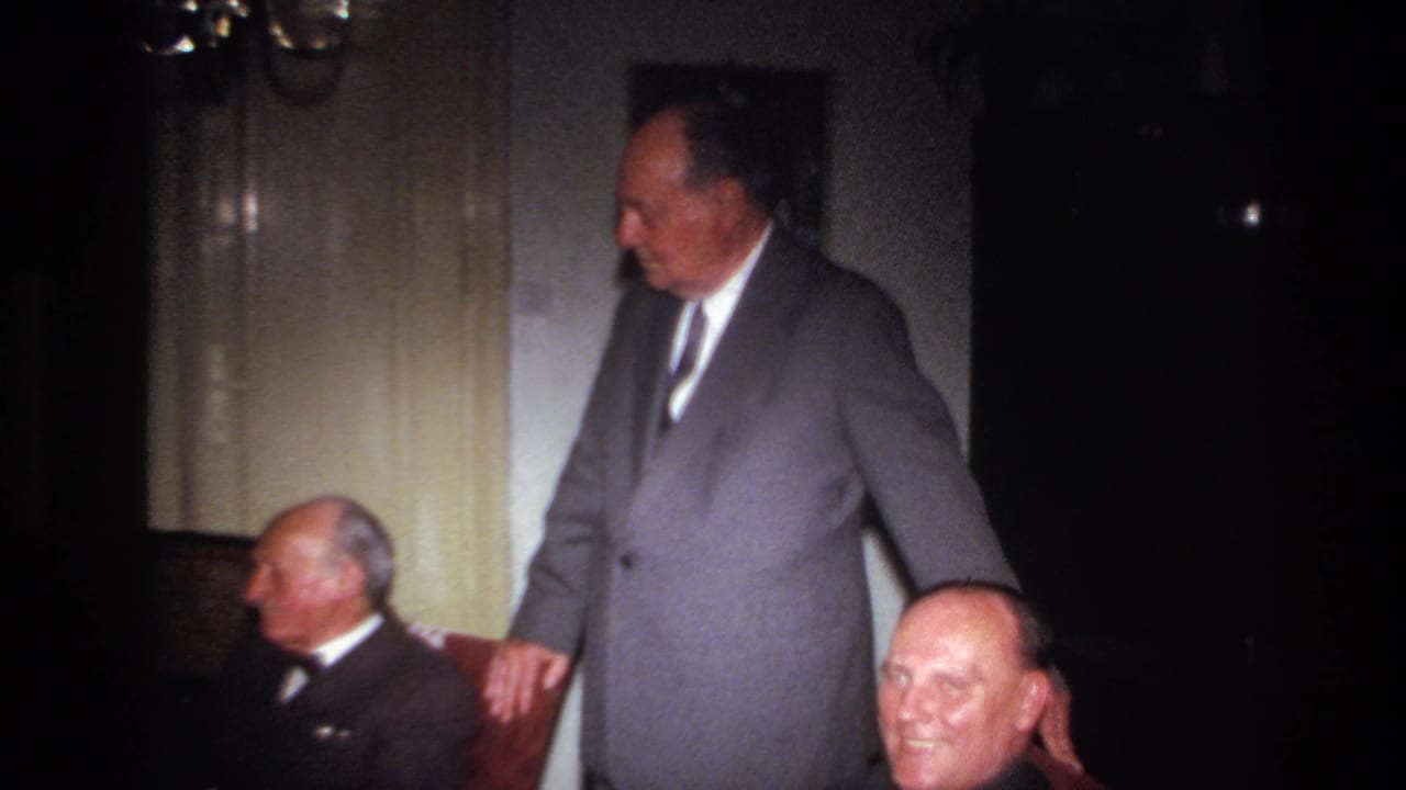 1967 French home-movie frame showing adult men gathered indoors, with one standing by a chair and others seated nearby.