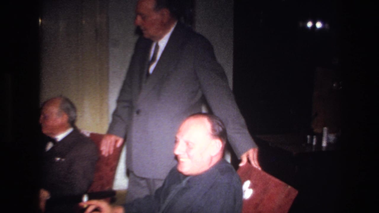1967 France indoor gathering with a suited man standing beside a chair and a seated man smiling toward the camera.