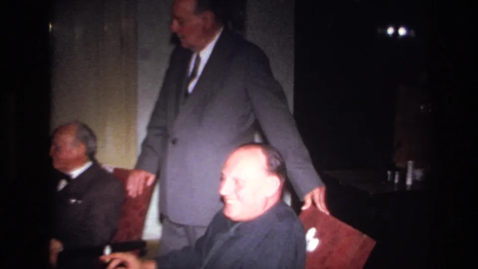 1967 France indoor gathering with a suited man standing beside a chair and a seated man smiling toward the camera.
