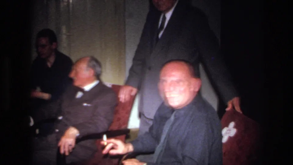 1967 French home-movie frame of a seated man holding a cigarette and smiling, with other adult men in the room.