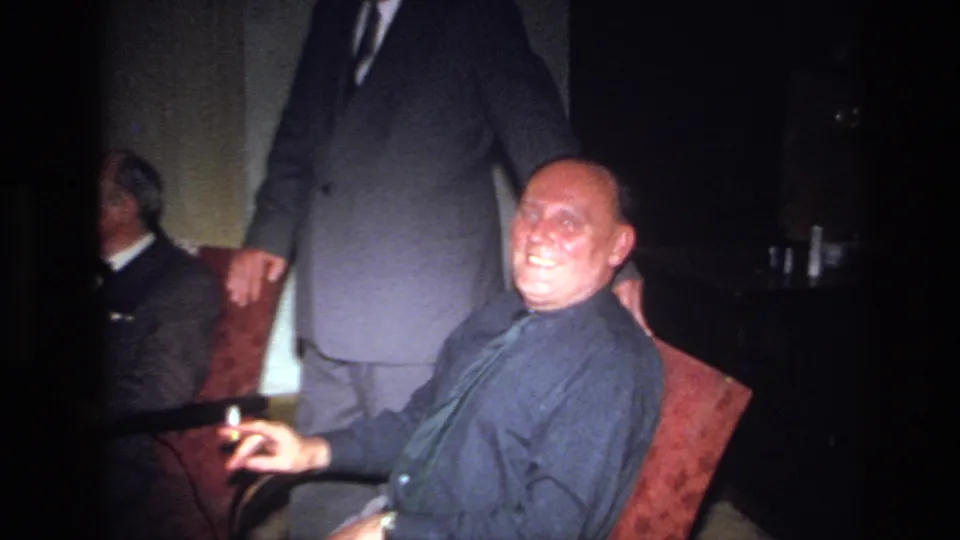 1967 France interior scene with a seated man smiling and holding a cigarette while others gather behind him.