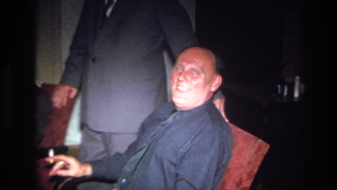 1967 France home-movie frame of a bald man in a dark shirt and tie smiling from a red chair as a suited man stands behind him.